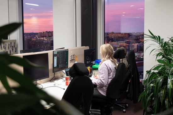 En medarbetare på qte sitter vid sitt skrivbord med rosafärgad solnedgång utanför fönstret.